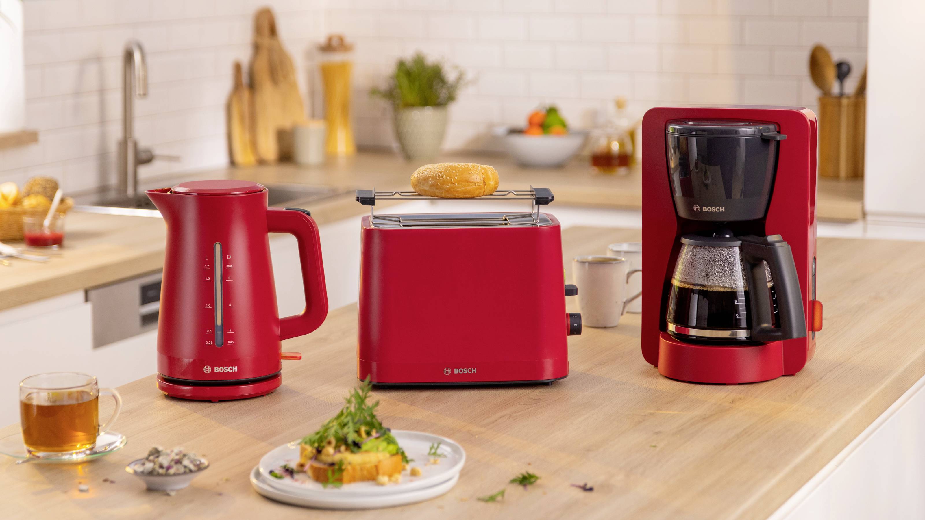 Un piano di lavoro della cucina con un bollitore elettrico rosso, un tostapane con toast e una macchina da caffè. Nelle vicinanze, un piatto con del cibo e una tazza da tè.