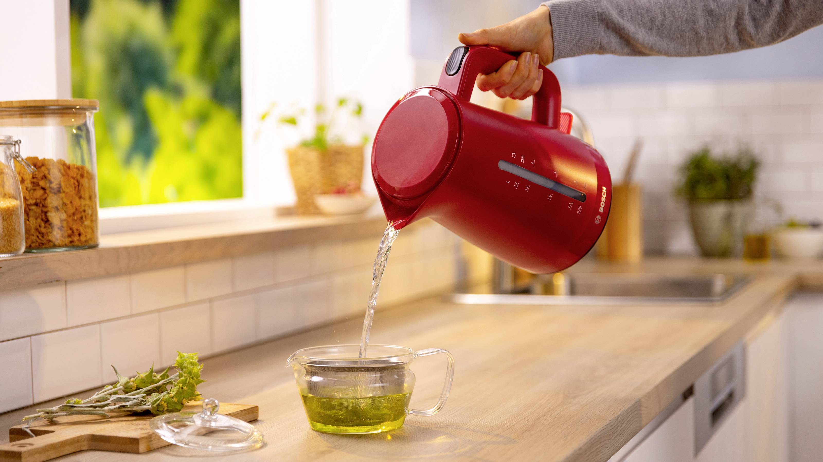 Una persona versa acqua calda da un bollitore elettrico rosso in una tazza di vetro con un infusore da tè su un piano da cucina, vicino a una finestra.