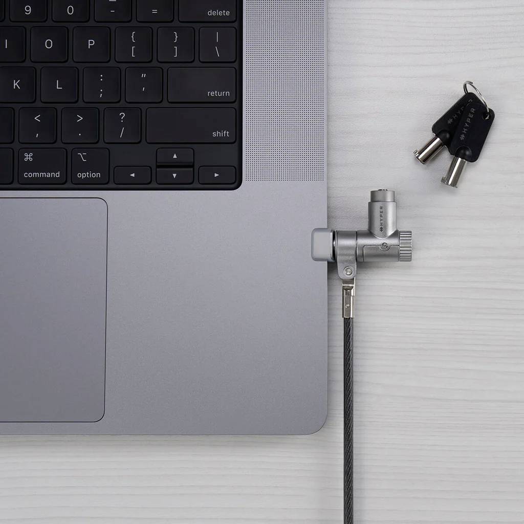 Un laptop con un lucchetto di sicurezza per cavo collegato sul lato destro, affiancato da due chiavi su un tavolo grigio chiaro, che illustra l'utilizzo sicuro di un dispositivo.