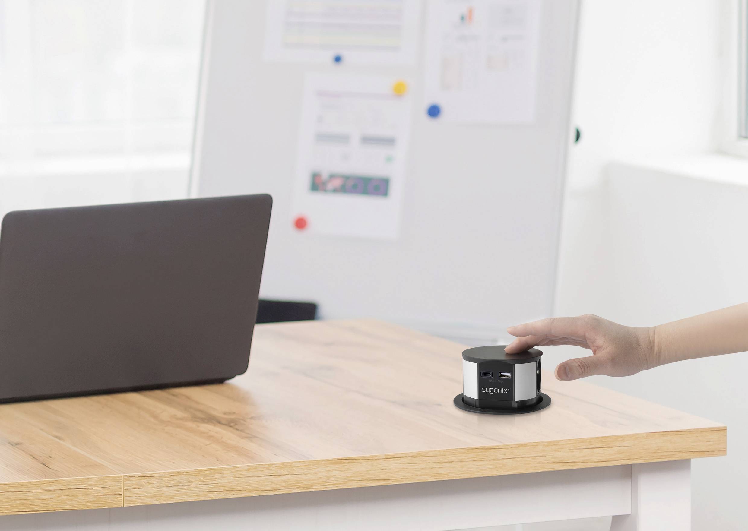 Una mano preme un dispositivo nero su un tavolo di legno accanto a un laptop, con una lavagna bianca e dei fogli sfocati sullo sfondo.