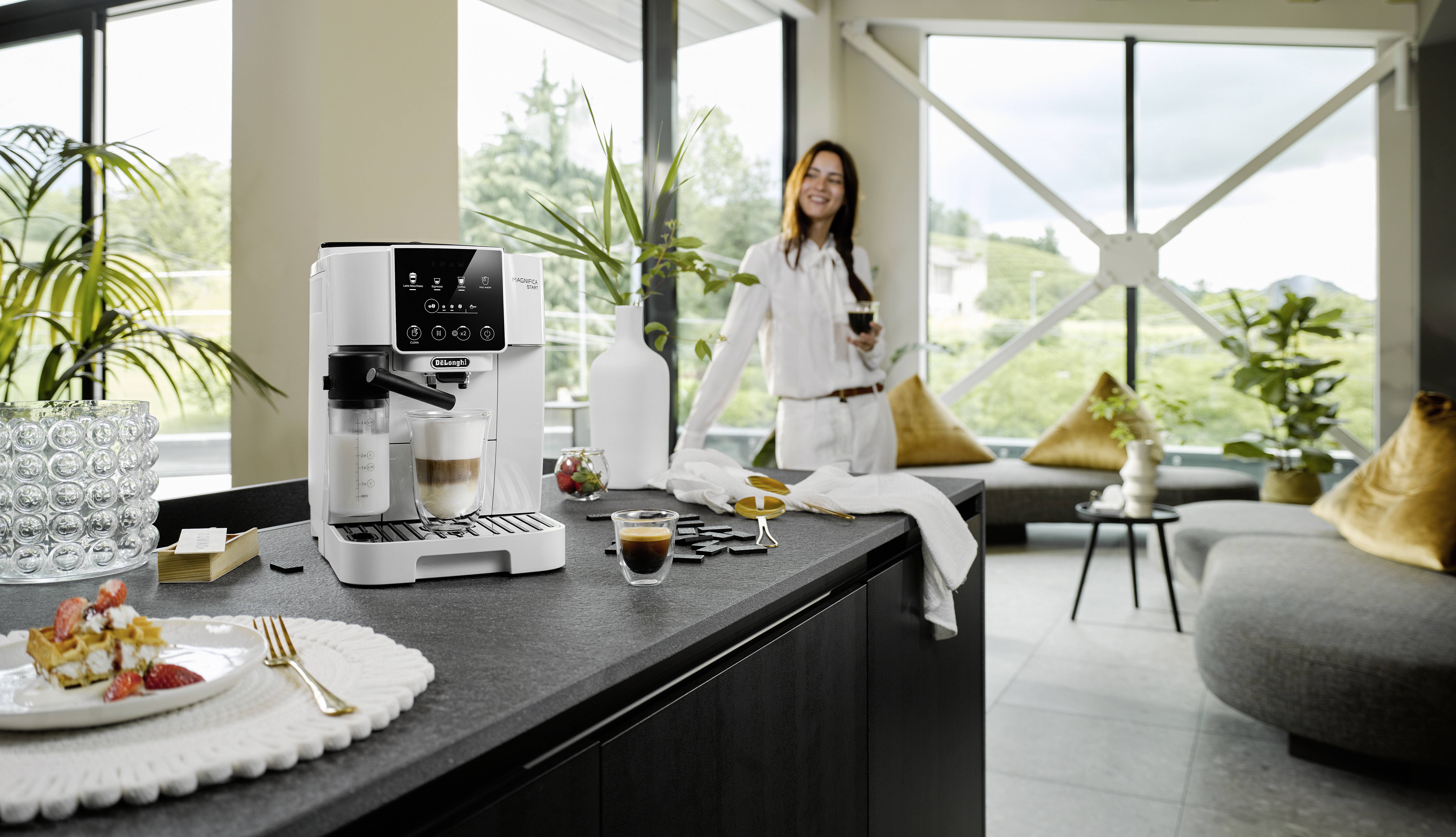 Una cucina moderna con una macchina da caffè sul bancone, accanto a dolci e bevande. Una persona sullo sfondo che tiene in mano una tazza di caffè.