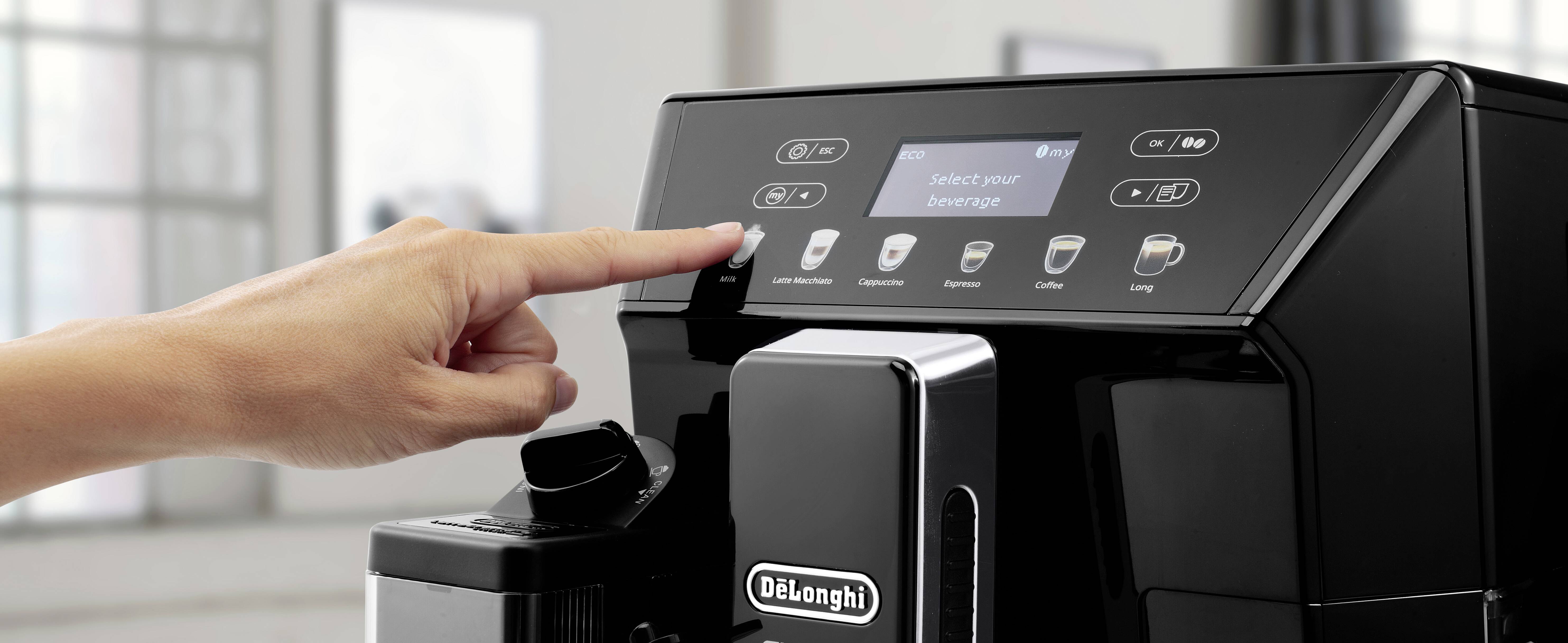 Una persona preme un pulsante sullo schermo di una macchina da caffè De'Longhi che mostra 'Seleziona la tua bevanda' in un ambiente cucina moderno.
