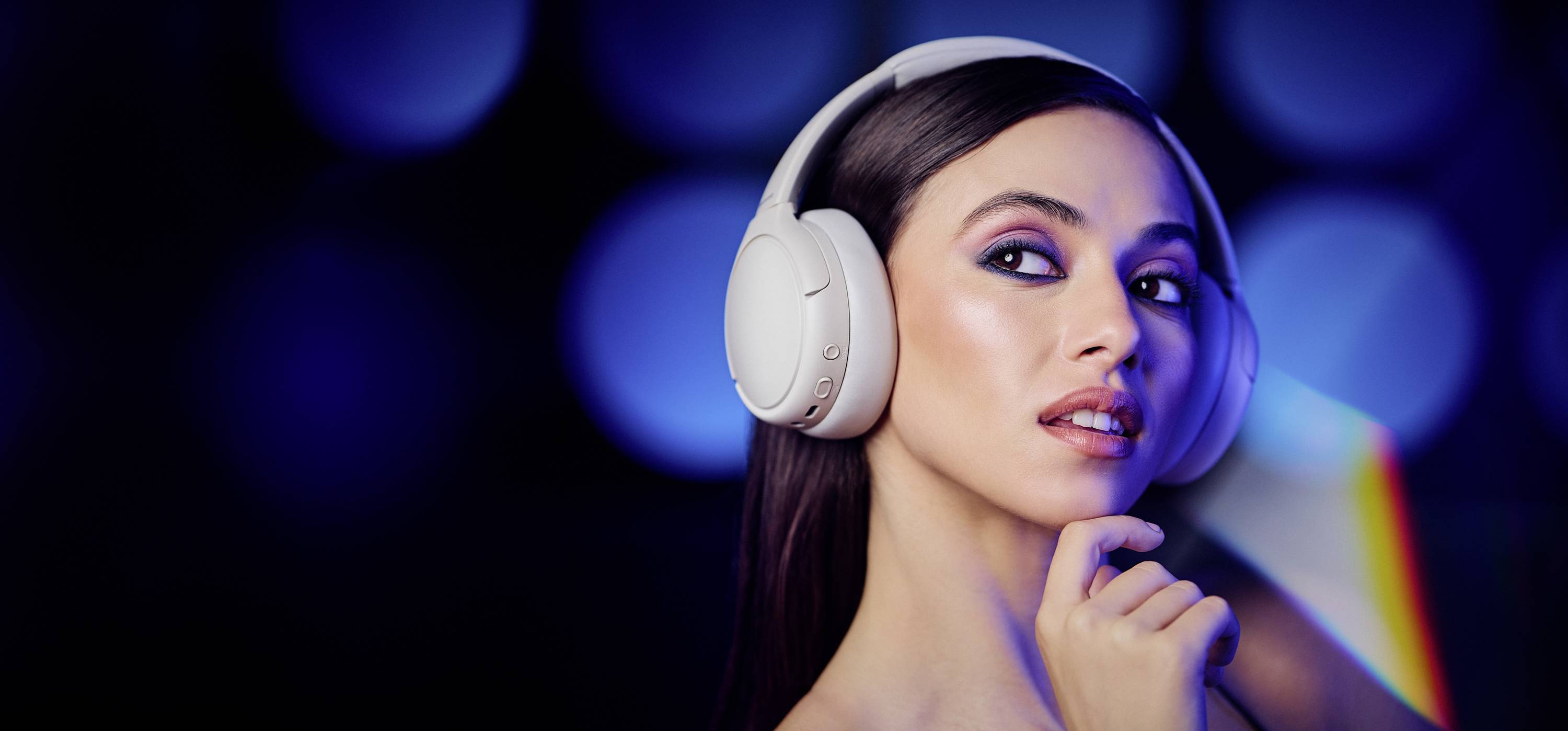 Una donna con cuffie bianche è in piedi davanti a uno sfondo sfocato e multicolore. Guarda oltre la sua spalla con un'espressione pensierosa.