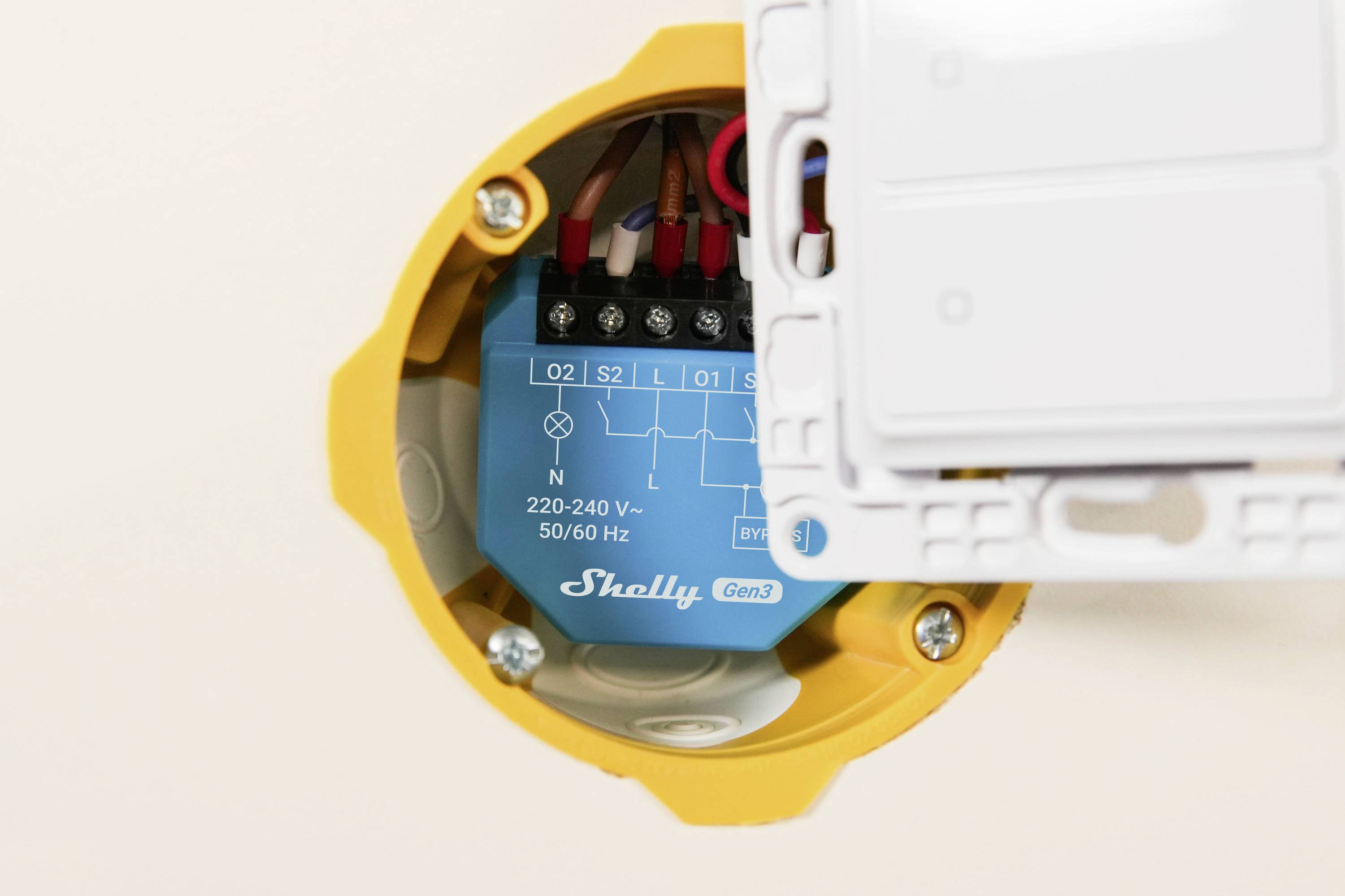 Un primo piano di un dispositivo elettrico montato in un alloggiamento circolare giallo. Il componente blu all'interno riporta 'Shelly Gen3' con istruzioni di cablaggio.