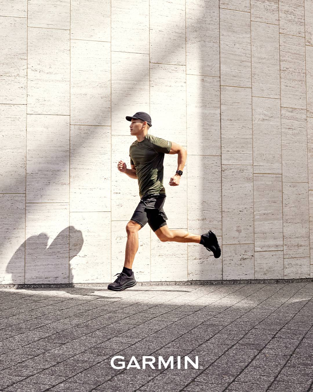 Un uomo in abbigliamento sportivo corre lungo un muro assolato e con una texture. Le ombre proiettano un pattern dinamico. Il nome del brand 'Garmin' appare sotto.