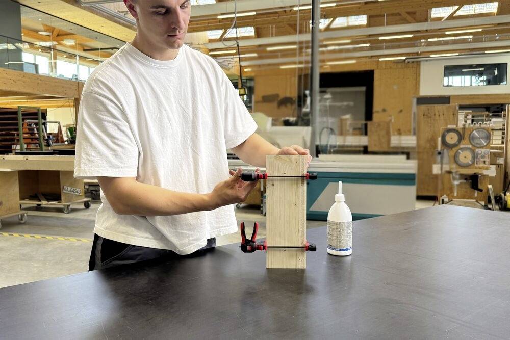 Una persona in un laboratorio che assembla un pezzo di legno utilizzando morsetti e colla, circondato da utensili e attrezzature per la lavorazione del legno.