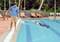 Una persona usa una rete per la pulizia della piscina con un manico estensibile accanto a una grande piscina. Diversi lettini sono sullo sfondo.