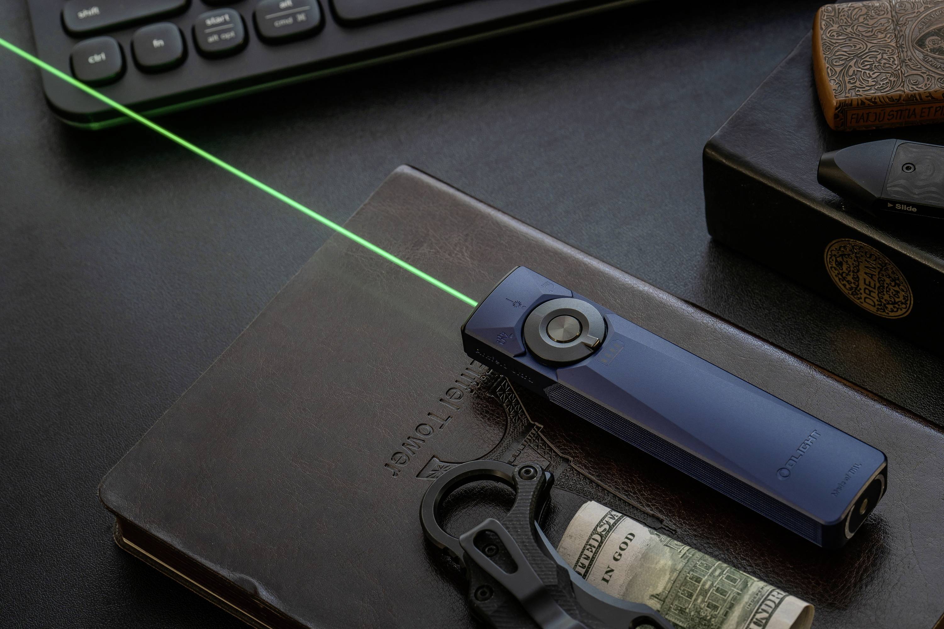 Un puntatore laser blu proietta un fascio verde su una scrivania con libri chiusi, una tastiera, un multiutensile e un rotolo di banconote nelle vicinanze.