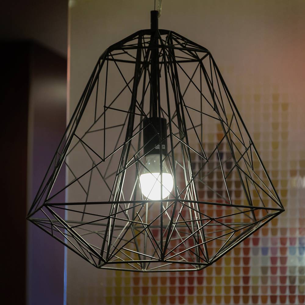 Un pendente luminoso con struttura geometrica a wireframe, con una lampadina che emana luce, è appeso contro una parete pixelata dai colori vivaci.