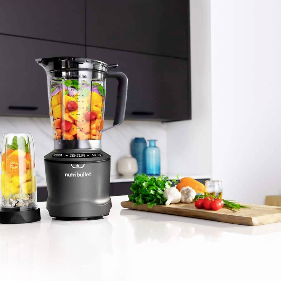 Un frullatore NutriBullet nero e un'alta tazza abbinata sono riempiti di frutta e verdura colorata. Sono posizionati su un piano di lavoro della cucina.