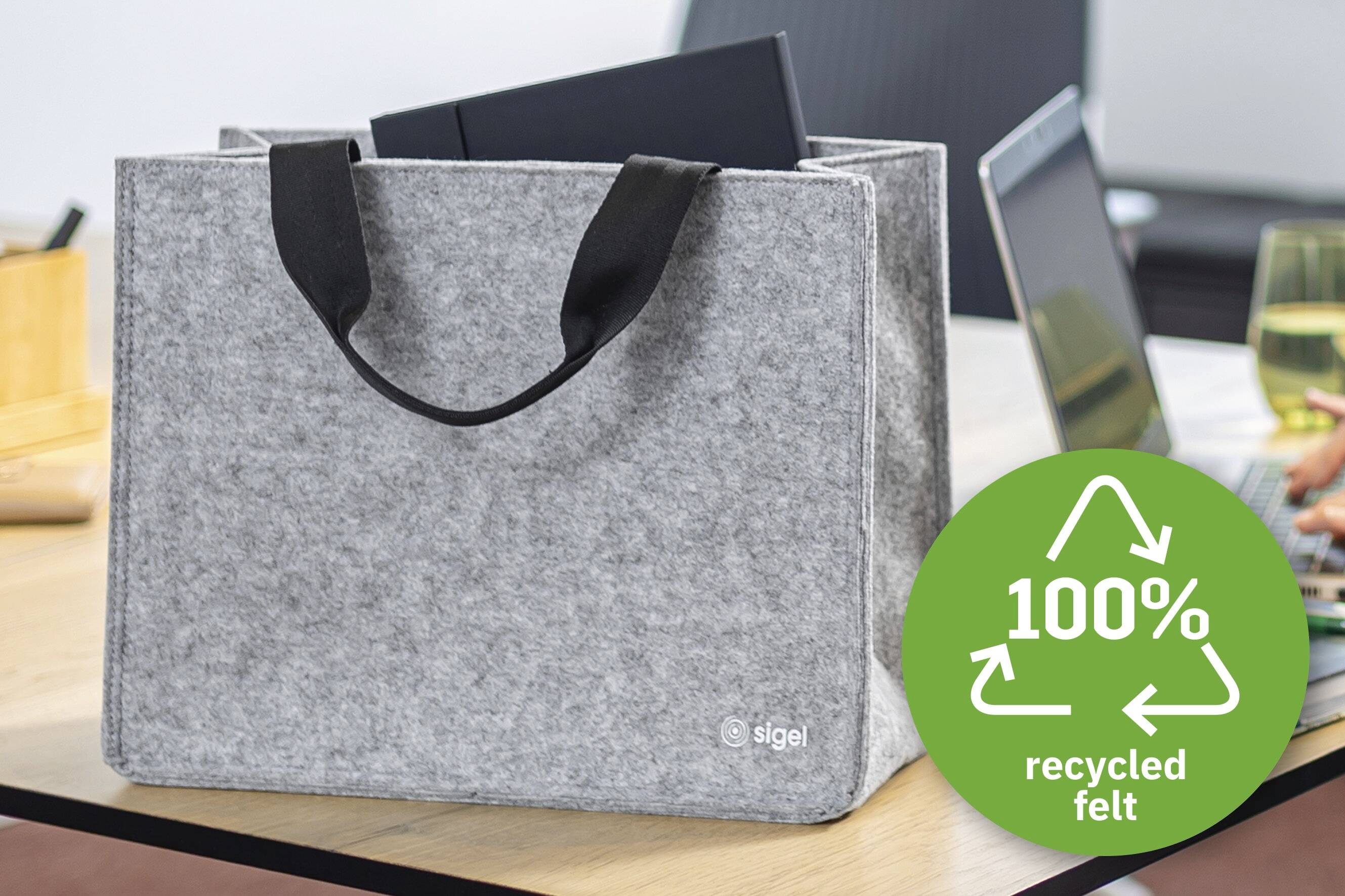 Una borsa a tracolla grigia in feltro con manici neri su un tavolo, etichettata 'sigel'. Un simbolo circolare verde riporta '100% feltro riciclato'.