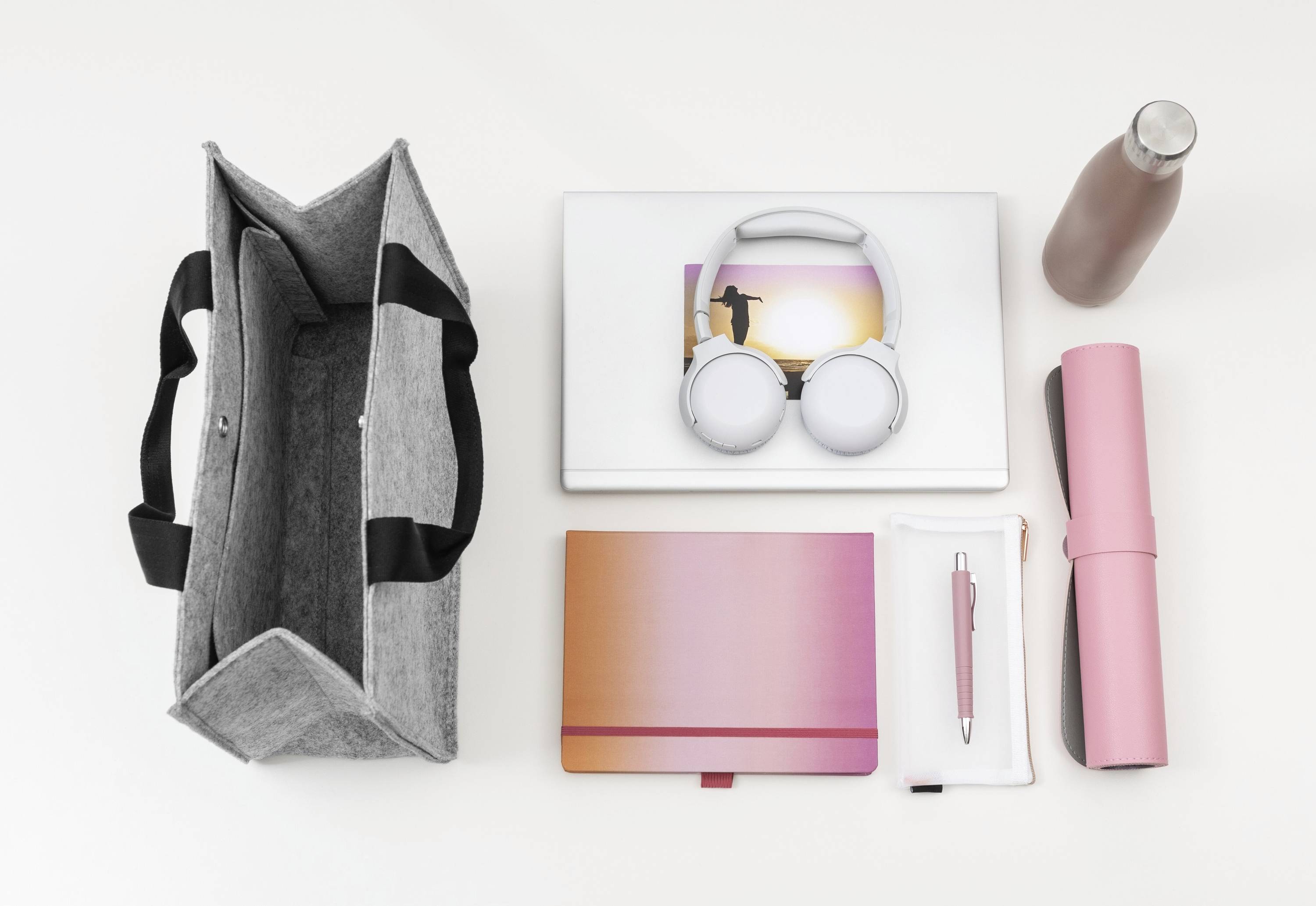 Borsa a tracolla grigia, laptop con cuffie, bottiglia d'acqua rosa, quaderno, astuccio per penne e tappetino rosa ordinatamente disposti su una superficie bianca.