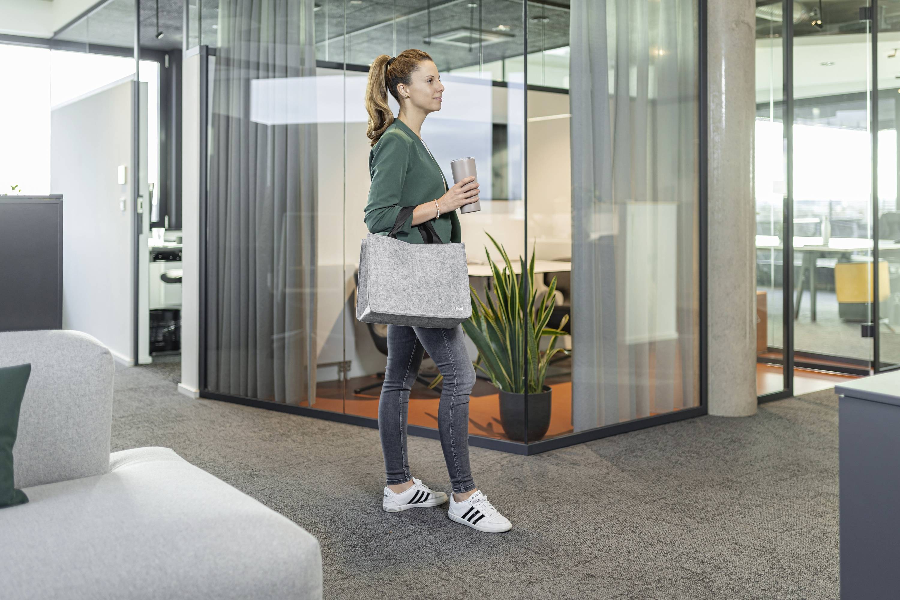 Una donna con jeans e una giacca verde cammina attraverso un ufficio moderno, tenendo in mano un tumbler e una borsa grigia, con pareti di vetro e una pianta nelle vicinanze.