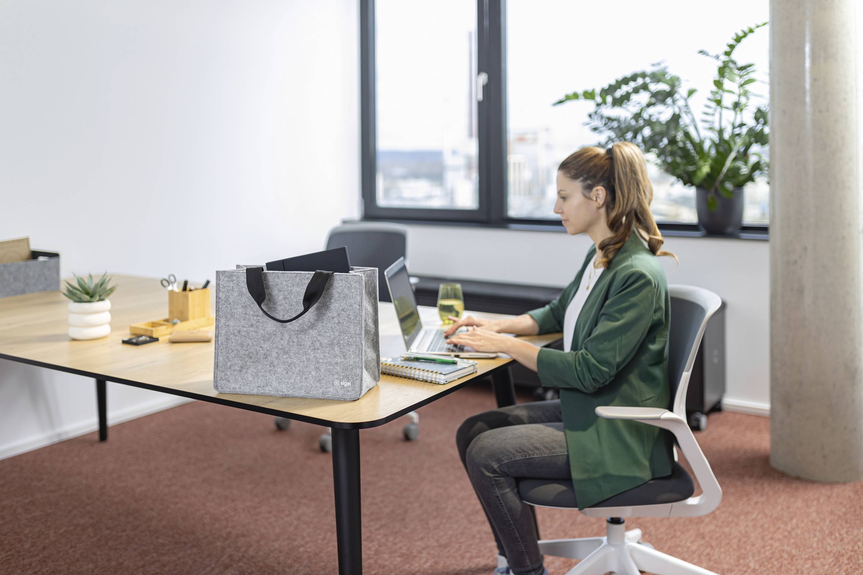 Una donna è seduta a una scrivania, digitando su un laptop in un ufficio moderno con una grande finestra, piante e una borsa di feltro sulla scrivania.