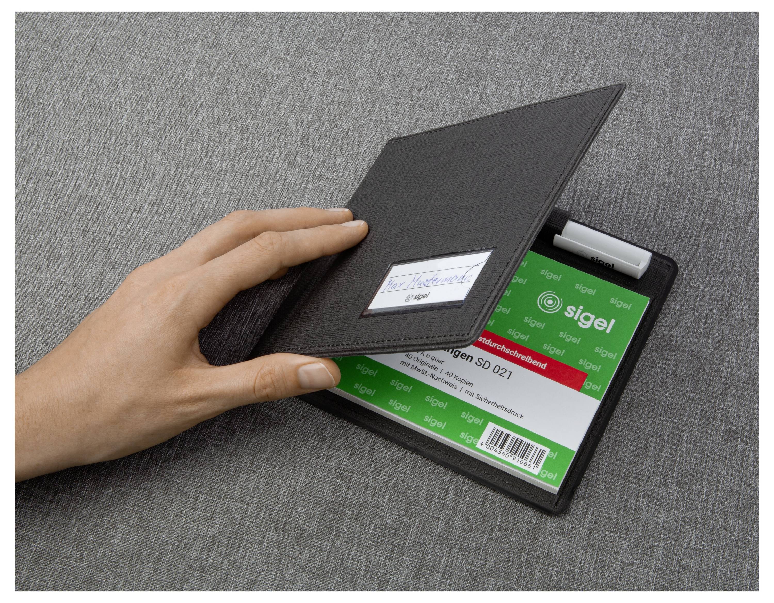Una mano che apre un portacarte da visita in stile quaderno nero mostrando una carta 'sigel' verde e una penna su una superficie grigia.