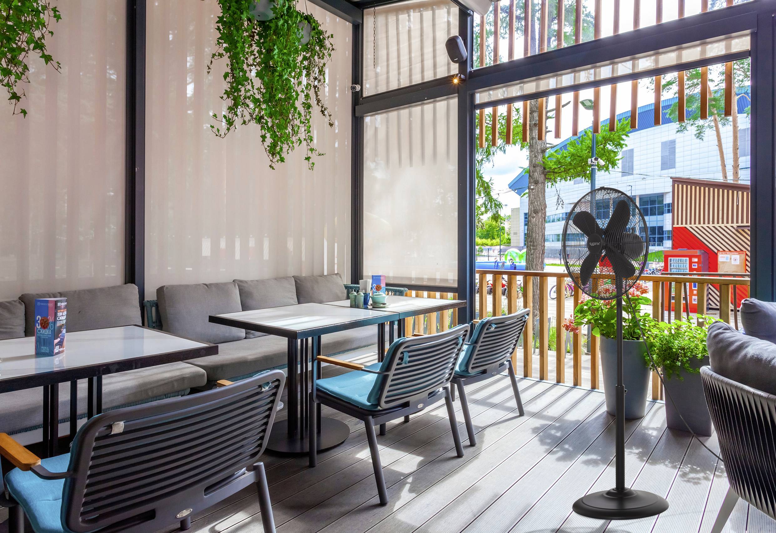 Un patio di un caffè all'aperto con tavoli e sedie, circondato da piante. Un ventilatore è nell'angolo, e gli edifici sono visibili sullo sfondo.