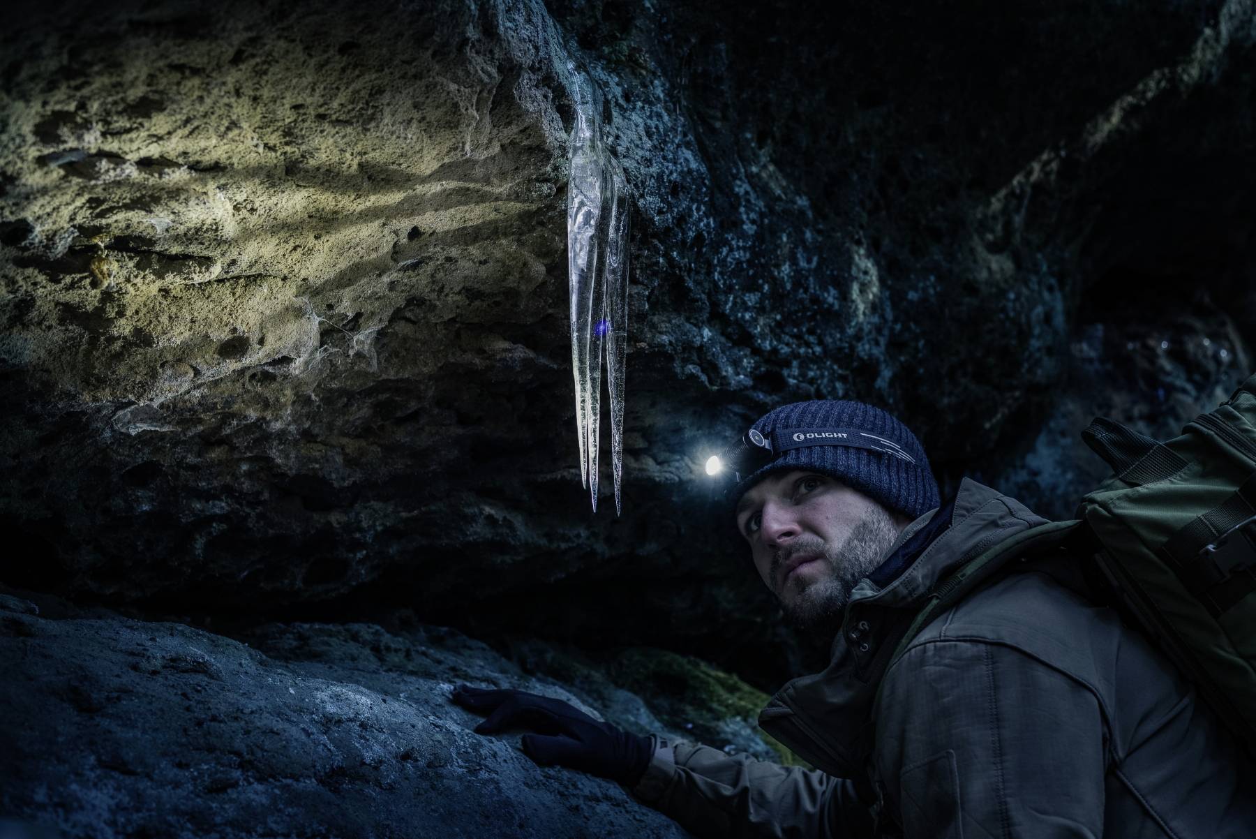 Una persona con un cappotto invernale e una lampada frontale osserva i ghiaccioli appesi al soffitto di una grotta buia, illuminati dalla luce della lampada frontale.