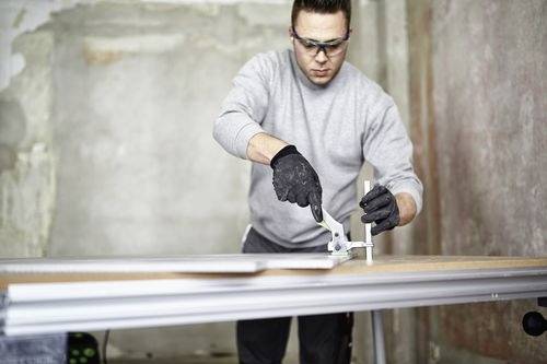 Una persona che indossa occhiali protettivi e guanti utilizza un taglierino su un pezzo di materiale, in un ambiente industriale o di officina.