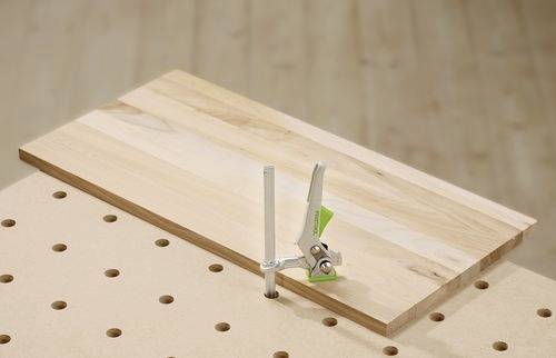 Un'asse di legno fissata su un banco da lavoro forato con una morsa metallica, utilizzata per lavori di falegnameria o carpenteria.
