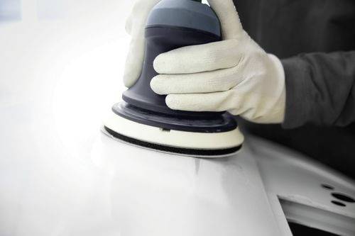 Una persona che indossa dei guanti utilizza una levigatrice elettrica su una superficie liscia e bianca di un'auto, suggerendo un processo di riparazione o manutenzione del veicolo.