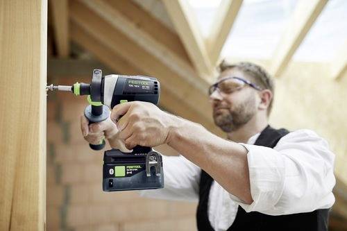 Un uomo con occhiali protettivi usa un trapano a batteria per avvitare una vite in una trave di legno, parte di un progetto di costruzione o falegnameria.
