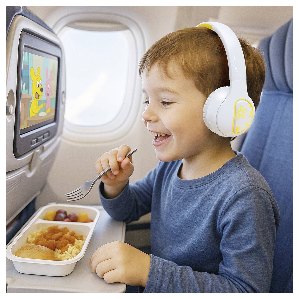 Un bambino su un aereo, con le cuffie, sorride mentre guarda un cartone animato sullo schermo del sedile. Sta mangiando un pasto da un vassoio.