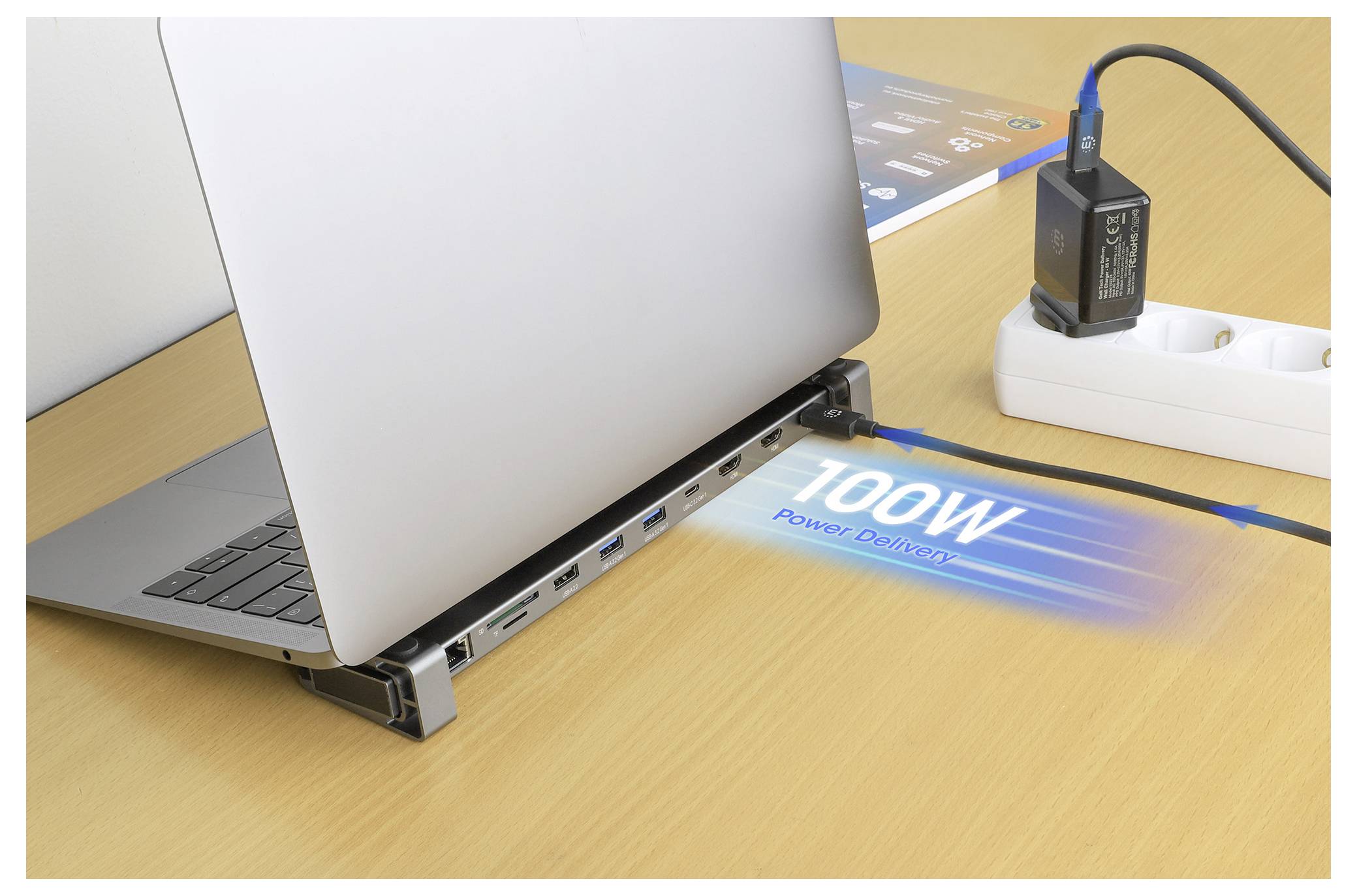 Un laptop poggia su una stazione di aggancio che proietta '100W Power Delivery' sulla scrivania, enfatizzando l'elevata capacità di carica. Una ciabatta elettrica con un adattatore inserito è visibile.