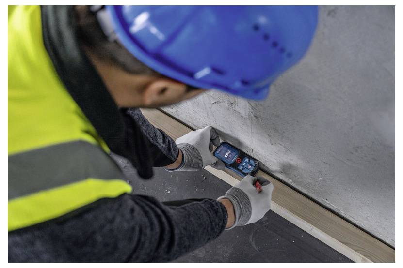 Un operaio edile con un elmetto blu e un giubbotto catarifrangente utilizza un misuratore di distanza laser su una parete, concentrandosi su misurazioni precise.