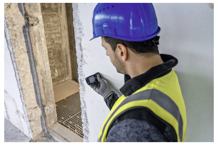 Un lavoratore edile con un casco blu e un giubbotto giallo utilizza un dispositivo portatile per misurare lo spessore di un muro in calcestruzzo in un cantiere edile.