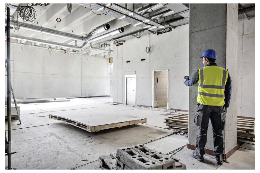 Operaio edile con giubbotto alta visibilità e casco valuta uno spazio interno vuoto in calcestruzzo, suggerendo lavori di ristrutturazione in corso.