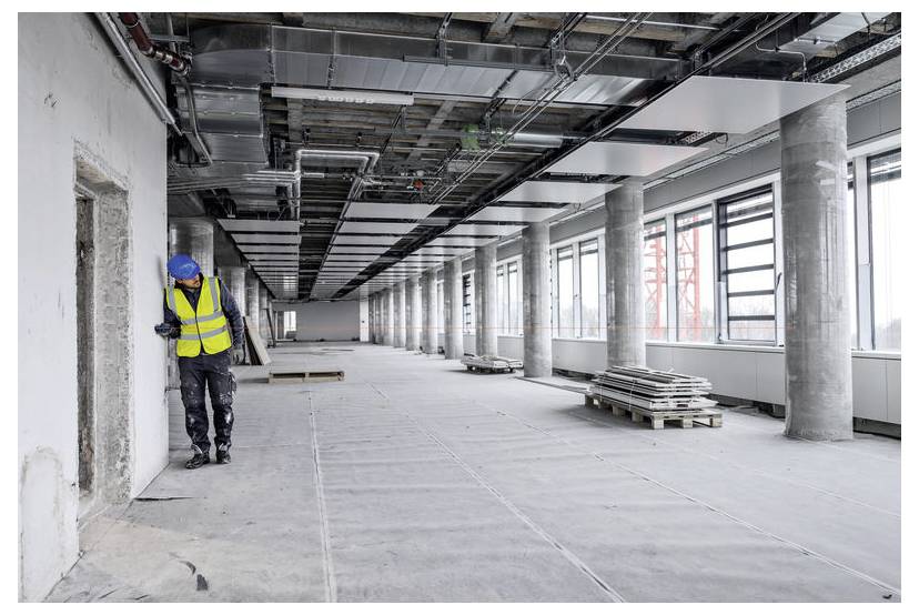 Un lavoratore edile con giubbotto di sicurezza e casco ispeziona un vano porta in un ampio spazio per uffici non ancora completato, con soffitti e colonne a vista.