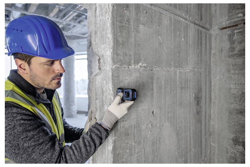Un operaio edile con un elmetto blu e un giubbotto di sicurezza utilizza uno strumento di misurazione su una parete in calcestruzzo all'interno di un edificio in costruzione.