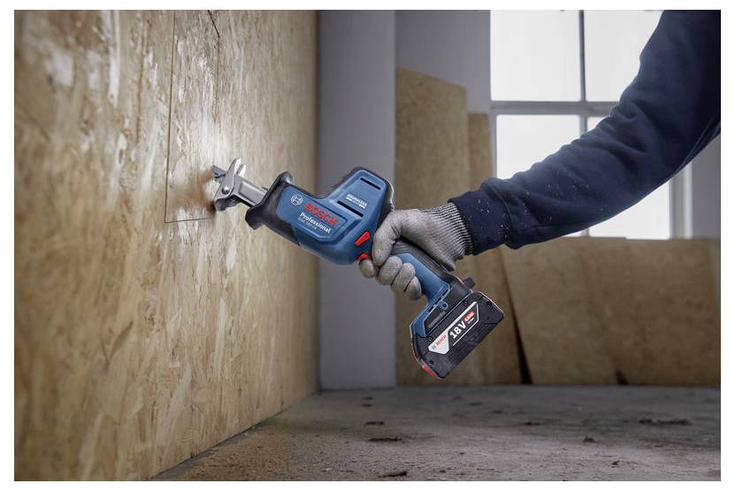 Una persona utilizza una sega a gattuccio cordless per tagliare un'asse di legno in un laboratorio. La finestra sullo sfondo illumina la scena.