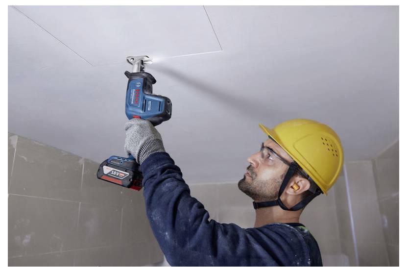 Un operaio edile che indossa un casco di sicurezza giallo e occhiali protettivi utilizza un trapano cordless per installare un pannello sul soffitto di una stanza.