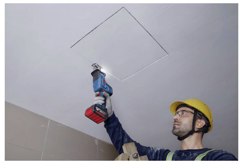Un lavoratore edile con dispositivi di sicurezza utilizza un utensile elettrico per tagliare un'apertura quadrata nel soffitto, concentrandosi attentamente sul suo compito.