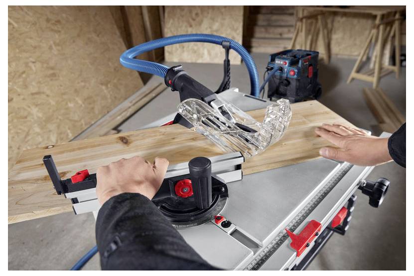 Una persona usa una sega circolare da tavolo per tagliare un'asse di legno in un laboratorio. La protezione di sicurezza e il tubo dell'aspiratore sono fissati alla sega.