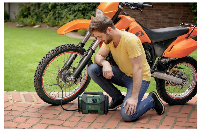 Un uomo è inginocchiato accanto a una moto arancione su un vialetto di mattoni, utilizzando un compressore portatile per gonfiare il pneumatico anteriore.