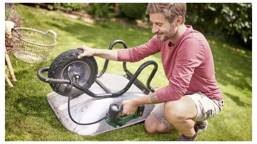 Una persona in un giardino ripara la ruota di un carrello ribaltato utilizzando un utensile elettrico. Giornata luminosa e soleggiata con erba verde e un cestello metallico nelle vicinanze.