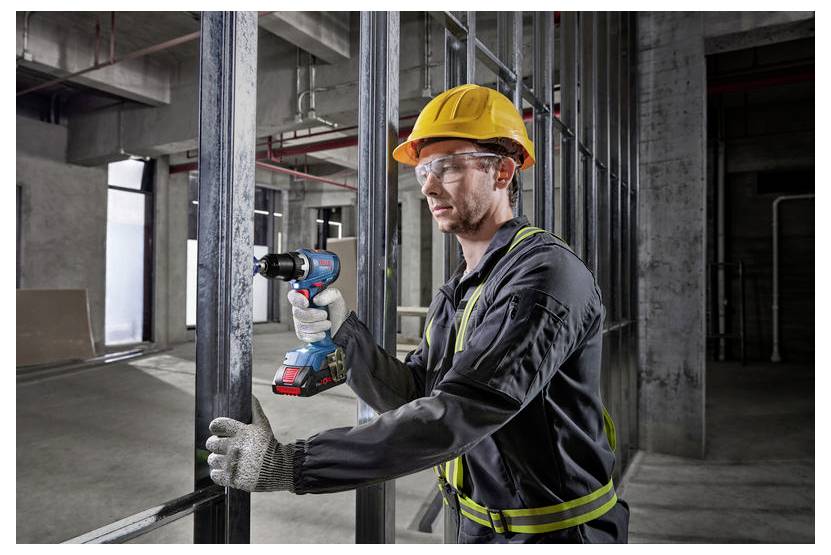 Un lavoratore edile con dispositivi di sicurezza, tra cui un elmetto e occhiali protettivi, usa un trapano elettrico su una struttura metallica in un edificio non ancora completato.