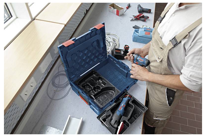 Una persona con tute da lavoro disimballa un trapano a batteria da una cassetta degli attrezzi su un tavolo da lavoro, circondato da vari utensili e materiali.