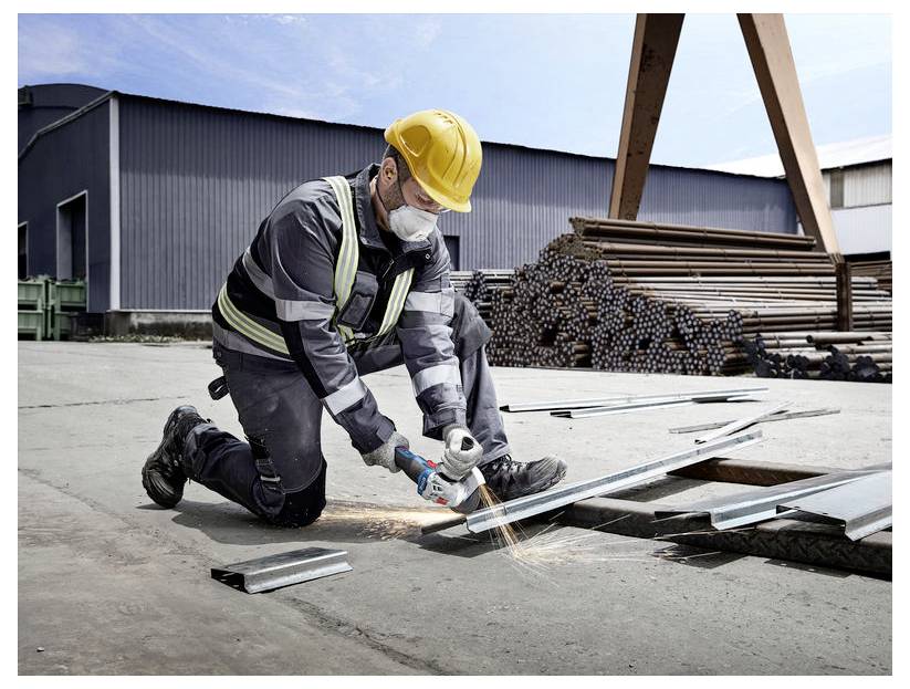 Un lavoratore edile è inginocchiato sul cemento, indossa un casco e dispositivi di sicurezza, utilizza un attrezzo elettrico per tagliare il metallo, con tubi impilati sullo sfondo.
