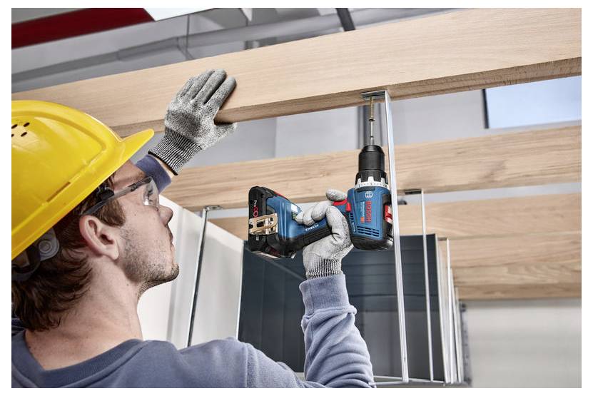 Persona che indossa un elmetto giallo utilizza un trapano cordless per fissare travi in legno, dimostrando pratiche di costruzione sicure.