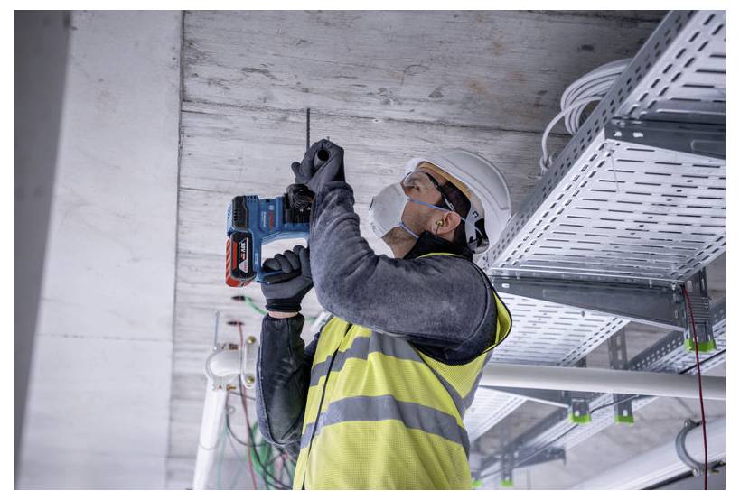 Un lavoratore edile con gilet di sicurezza e casco utilizza un trapano elettrico su un soffitto, circondato da cavi elettrici in un edificio non ancora completato.
