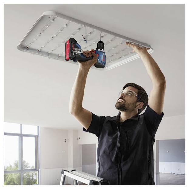 Una persona che indossa occhiali di sicurezza è in piedi su una scala e sta installando un grande pannello di illuminazione a soffitto utilizzando un trapano in una stanza luminosa.