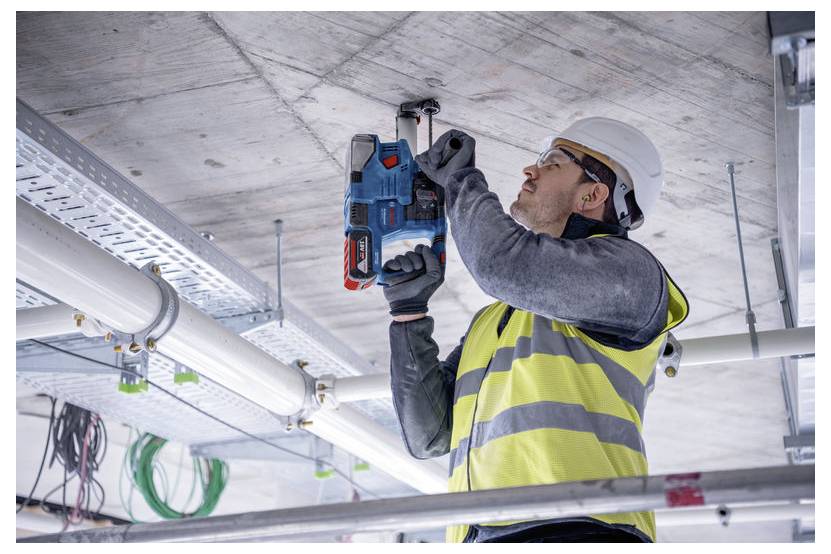 Un lavoratore edile con gilet di sicurezza e casco utilizza un utensile elettrico per forare un soffitto in calcestruzzo, circondato da tubi e cavi.