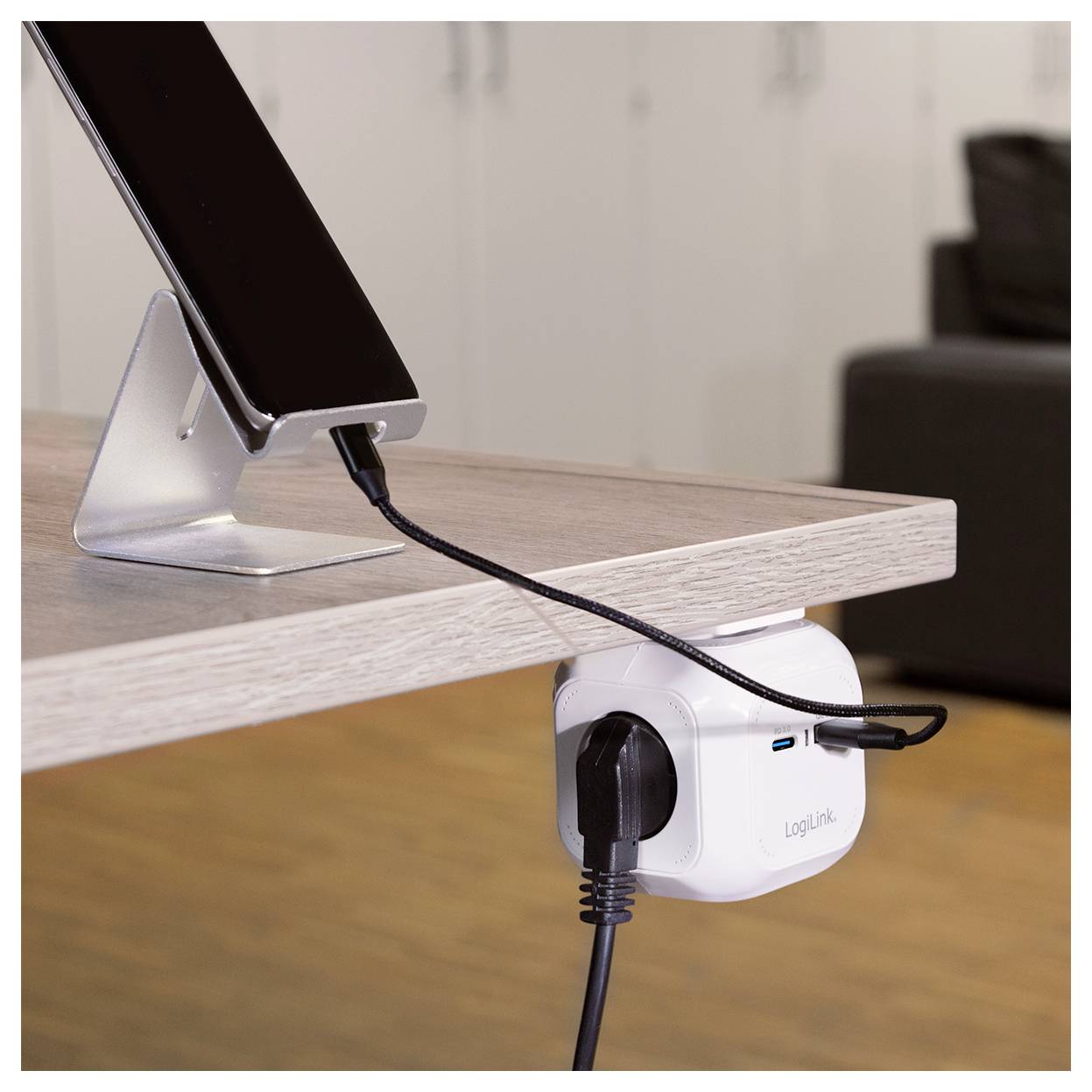 Un telefono su un supporto sta caricandosi tramite un cavo collegato a una presa elettrica montata sotto una scrivania in legno.