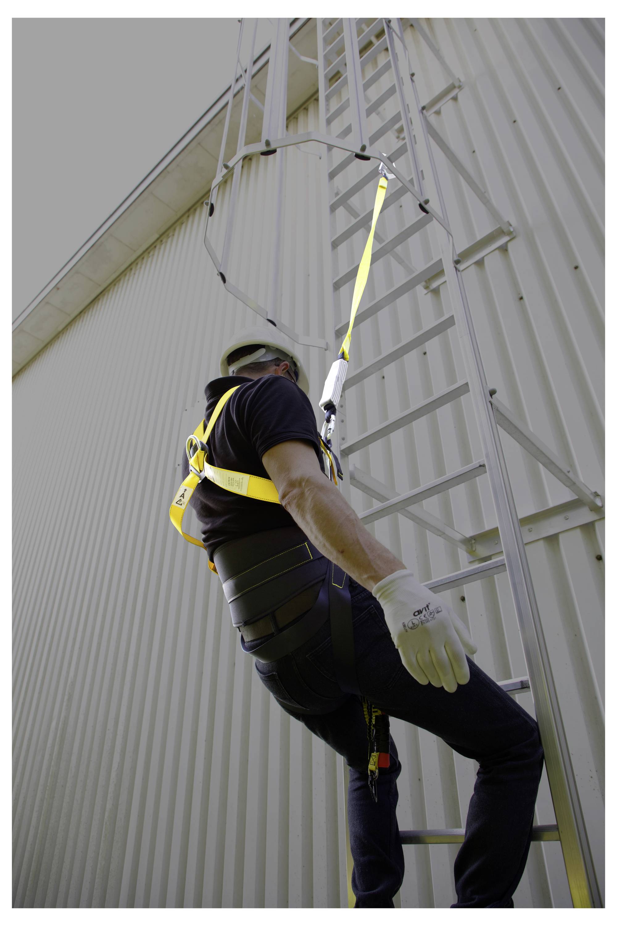 Lavoratore che indossa un'imbracatura di sicurezza sale una scala industriale sull'esterno di un edificio, evidenziando le precauzioni di sicurezza per il lavoro in quota.