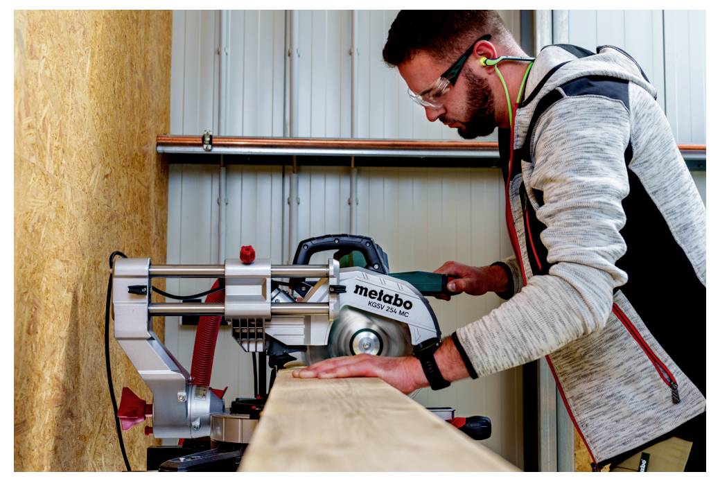 Una persona sta utilizzando una sega circolare per tagliare un'asse di legno in un laboratorio. Indossa occhiali protettivi e protezioni per l'udito.