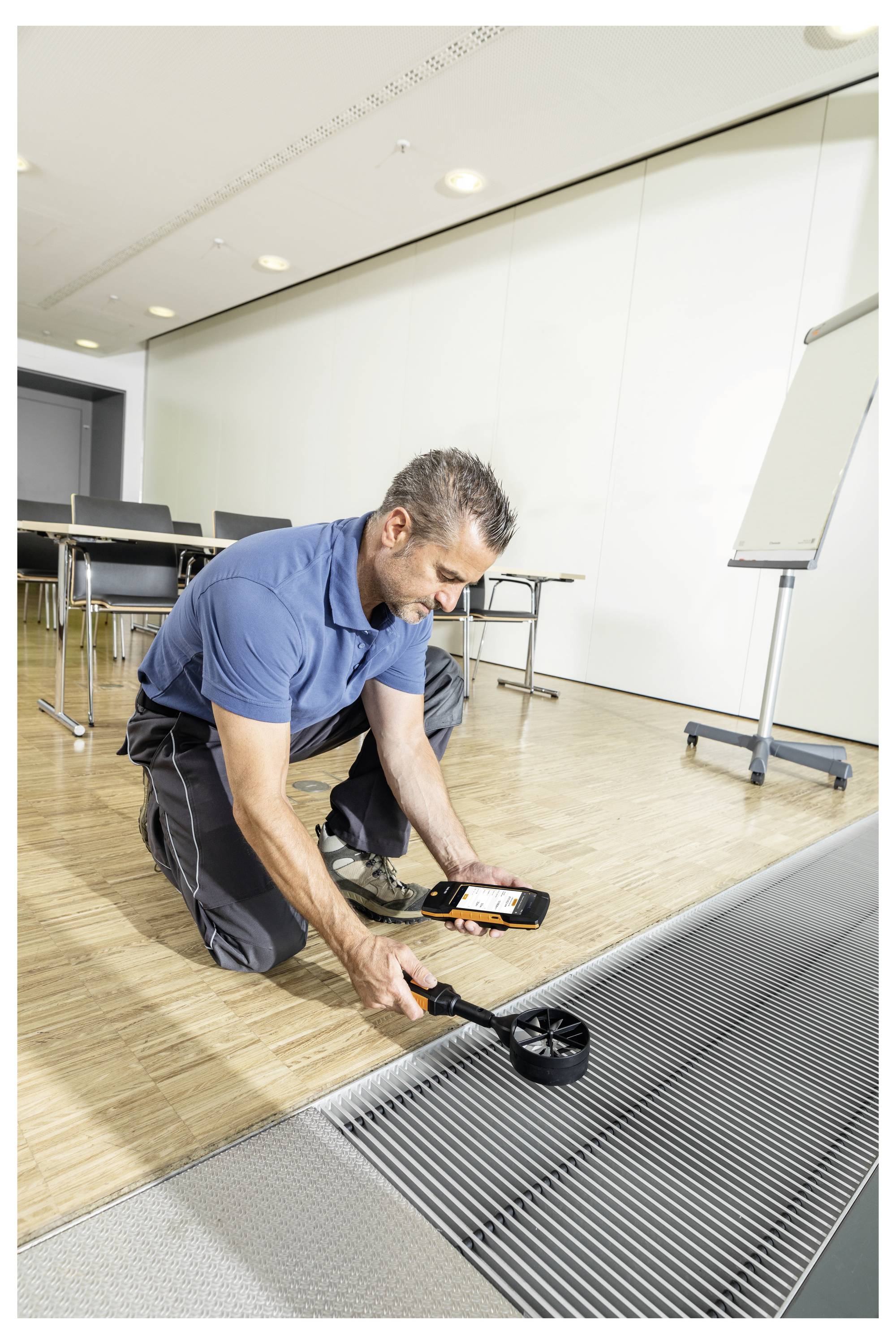 Un uomo è inginocchiato su un pavimento di legno in un ufficio, utilizzando un dispositivo portatile per misurare la qualità dell'aria vicino a una bocchetta di ventilazione metallica.