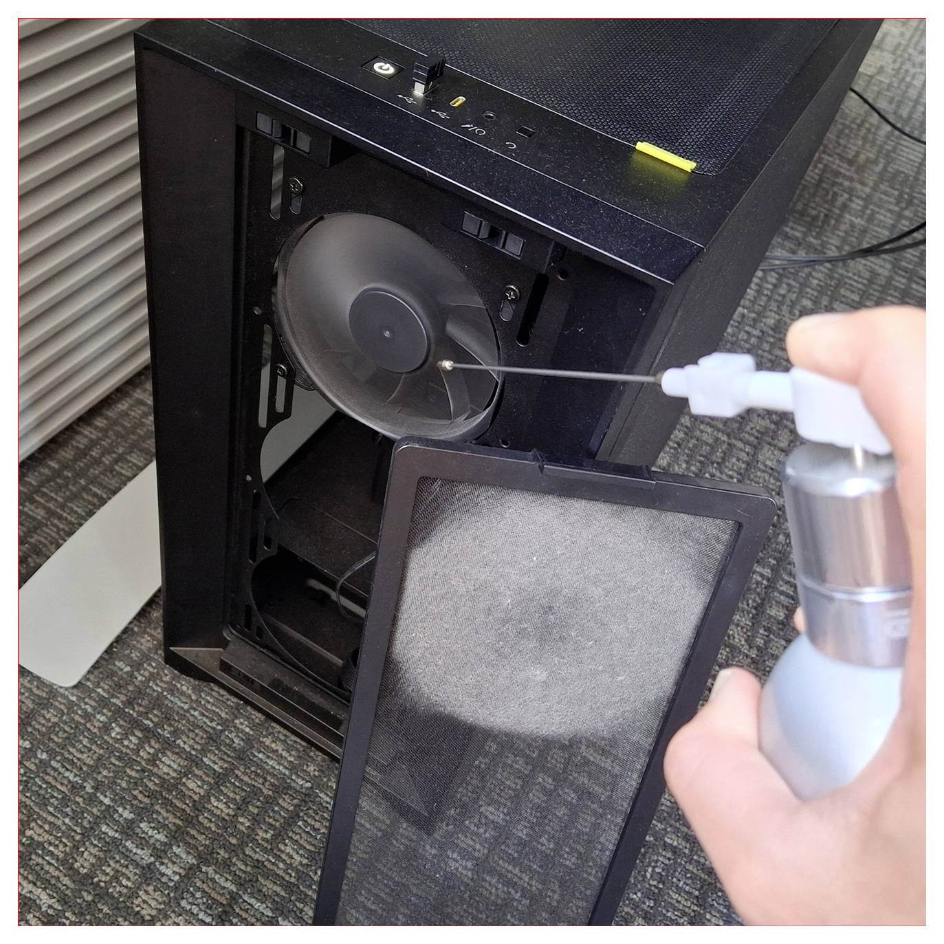 Una persona utilizza aria compressa per pulire un ventilatore del computer all'interno di un case torre con il filtro antipolvere rimosso sul pavimento.